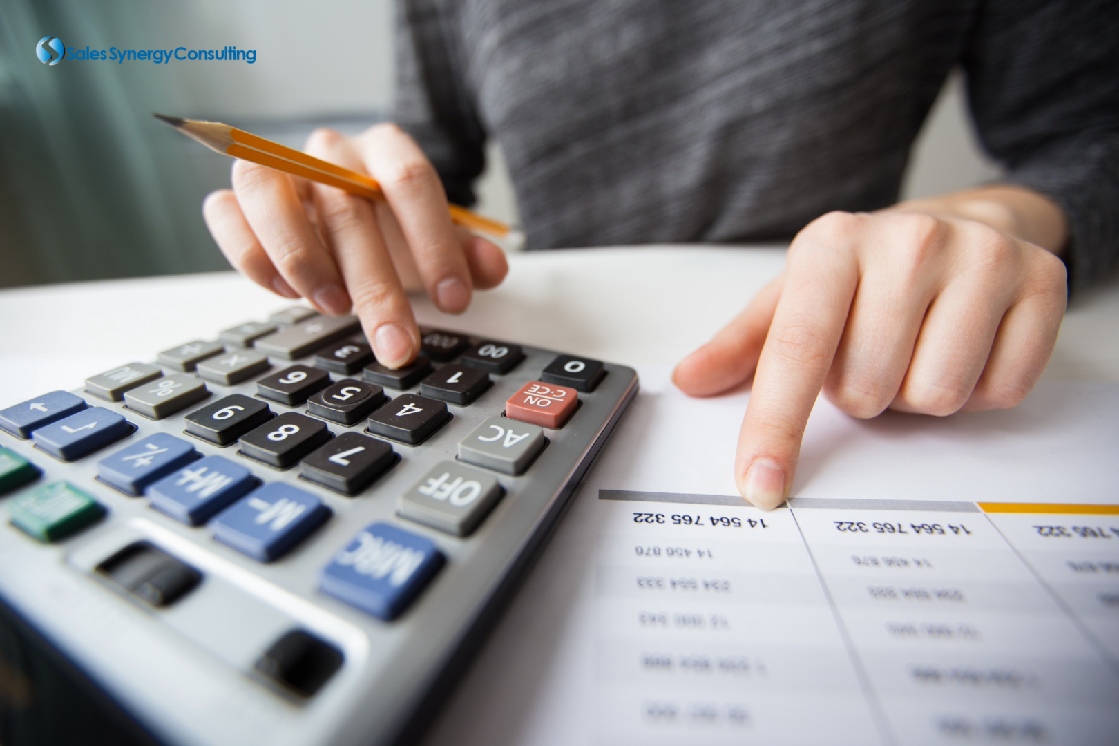 Person holding a pencil while typing into a calculator and pointing at a paper with numbers on it